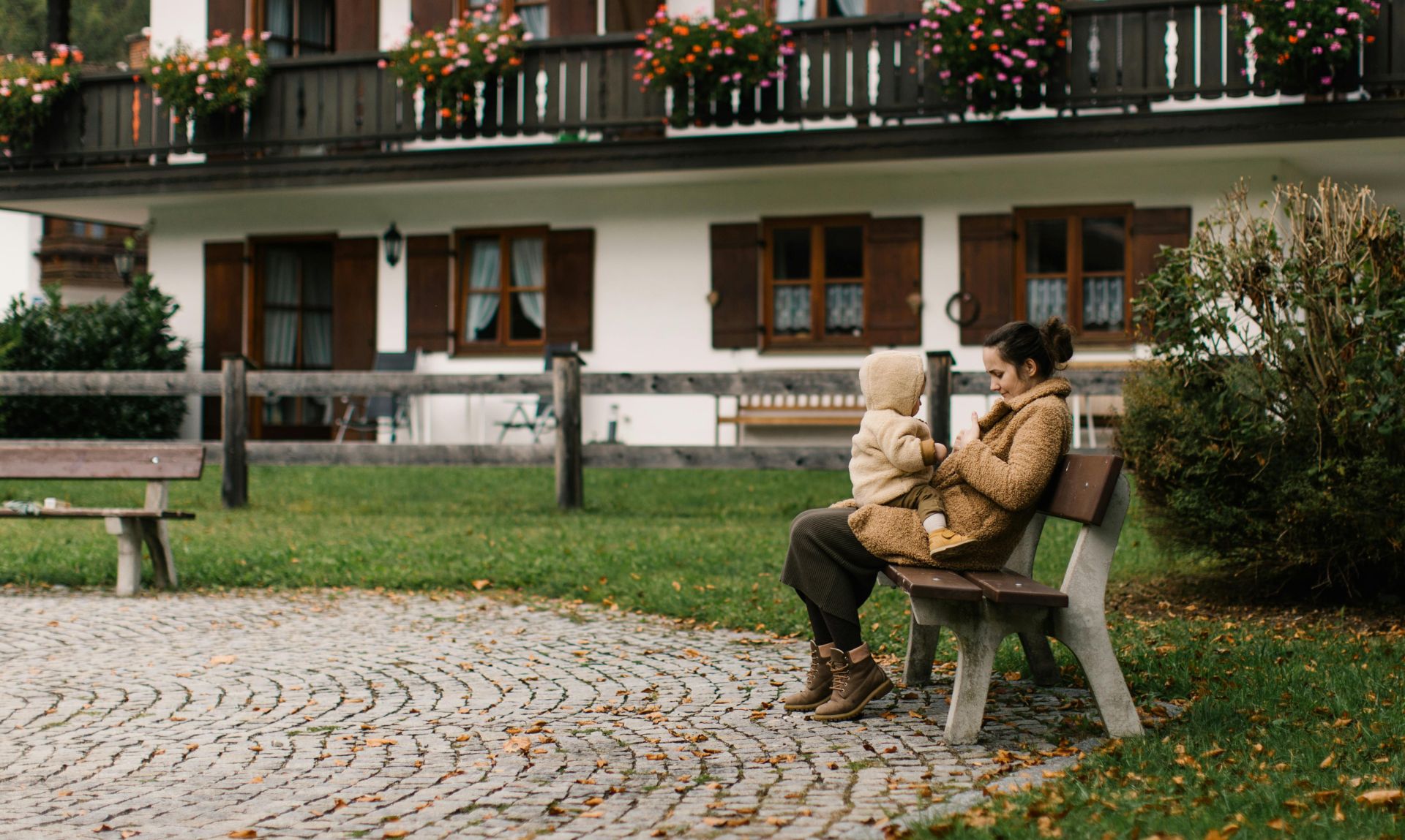 A mother and child in warm coats enjoy a moment on a bench outside a charming home with flowered balconies.